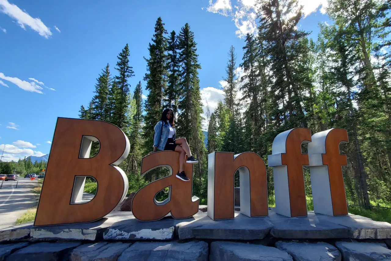 Person sitting on large 'Banff' sign with pine trees and blue sky in the background.
