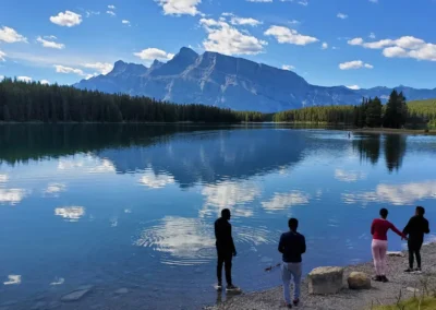 Two people stand by a serene lake, reflecting mountains and trees, while another walks nearby on the pebbled shore under a blue sky.