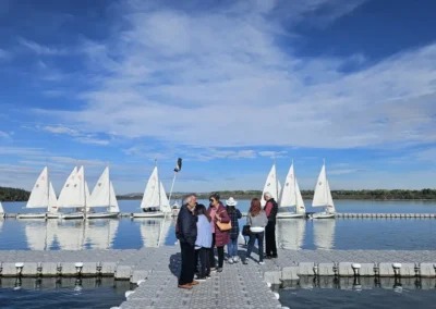A group of people converses on a dock with sailboats floating on a calm lake and a bright blue sky in the background.