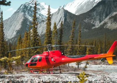 A bright red helicopter parked amidst green pine trees and rocky terrain, with majestic snow-capped mountains in the background.