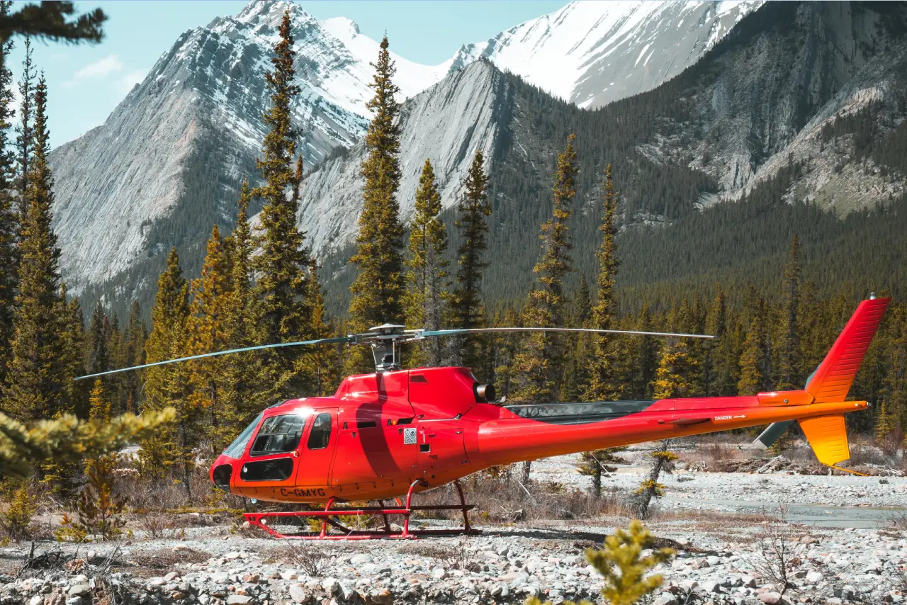 A bright red helicopter parked amidst green pine trees and rocky terrain, with majestic snow-capped mountains in the background.