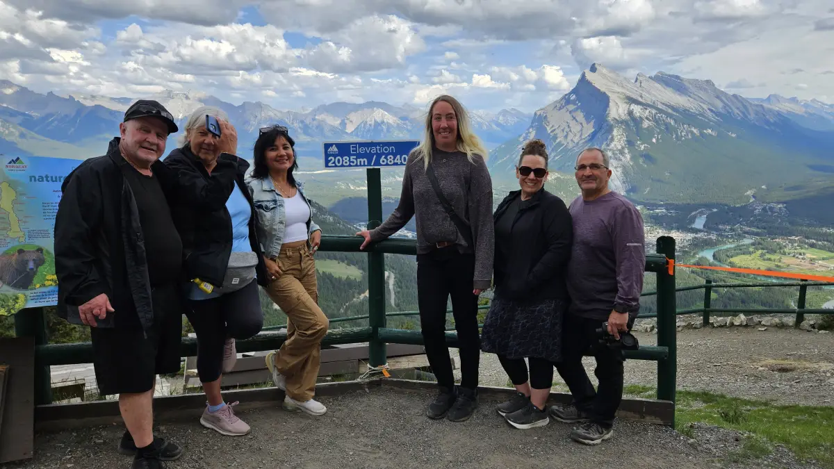 A group of six people poses at an overlook sign indicating 2085m elevation, with stunning mountains and valleys in the background.